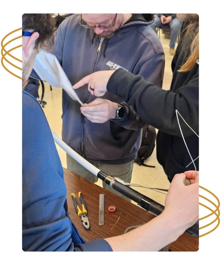 Three people assemble or repair an antenna with tools on a table, focusing on connecting parts.