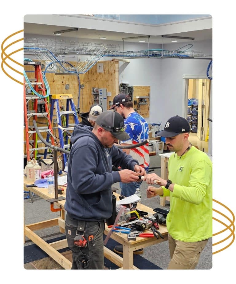 Two men work with tools in a workshop, while others work in the background among ladders and equipment.
