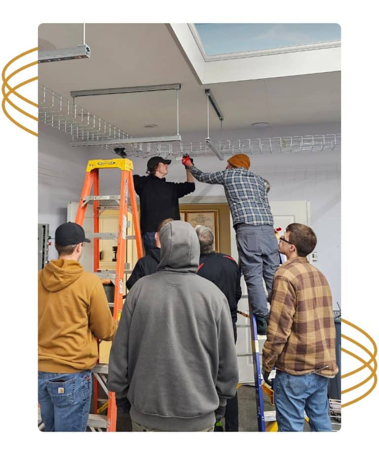 Six people watch as two others stand on ladders, installing a wire rack on the ceiling indoors.