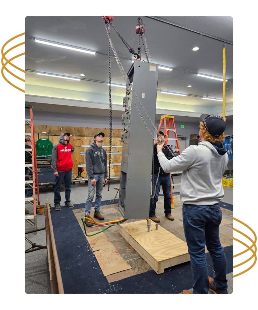 Four people use chains and pulleys to lift a large metal panel in an indoor workspace with ladders.